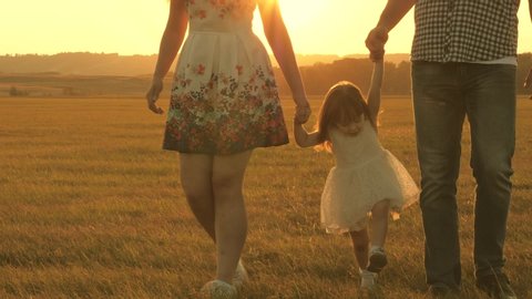 Walking with small kid in nature. little daughter jumping holding hands of dad and mom in park on background of sun. Family concept. child plays with dad and mom on field in sunset light.