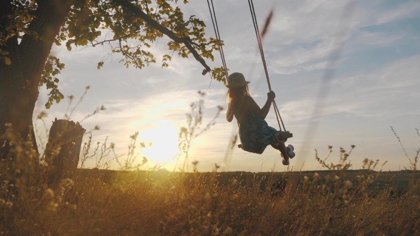 Happy child girl on swing at golden summer sunset. Silhouette of a young teenager girl swinging on the lone tree at nature, lifestyle. The concept of childhood dreams.