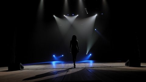 A popular young singer performs on stage in the light of bright flashing concert lighting, she goes on stage with a microphone in her hand. Slow motion.