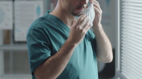 Tilt up shot of male hospital worker in scrubs and disposable hat putting medical mask on face and walking away