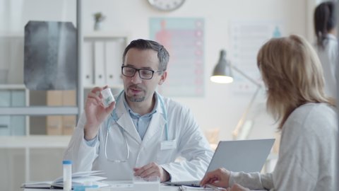 Caucasian male doctor in lab coat and glasses giving medicine to female patient, explaining instructions and writing prescription during consultation in clinic