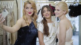 Portrait of three beautiful Caucasian fashion models in elegant dresses taking selfie. Camera approaching to young smiling women posing in boutique. Lifestyle, fashion, beauty, confidence. Slowmo.