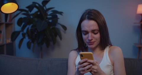 Close up of young girl crying while looking photos with her boyfriend after broke up. Sad young woman sitting on sofa looking at smartphone screen and weeping after receiving bad news .