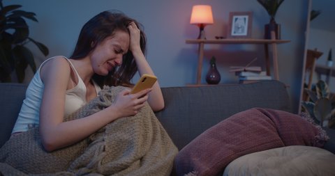 Depressed young woman sitting at sofa in tears. Upset caucasian girl broke up with boyfriend looking at smartphone screen and crying at home. People and Relationship Concept.