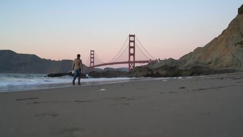Female Animal Lover Walking Playful Dog Along San Francisco's Baker Beach With Golden Gate Bridge In Background.