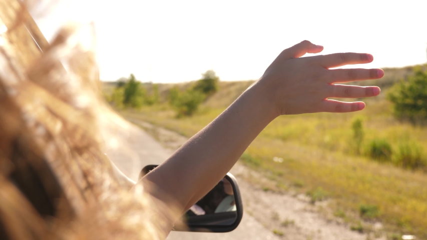 Free woman travels by car catches the wind with her hand from car window. Girl with long hair is sitting in front seat of car, stretching her arm out window and catching glare of setting sun