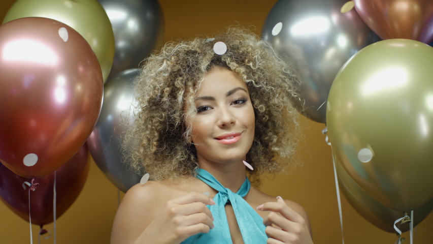 Young curly hispanic woman in turquoise dress dancing on yellow background with multicolored balloons and flying confetti. Pretty latin girl celebrates holiday at summer party