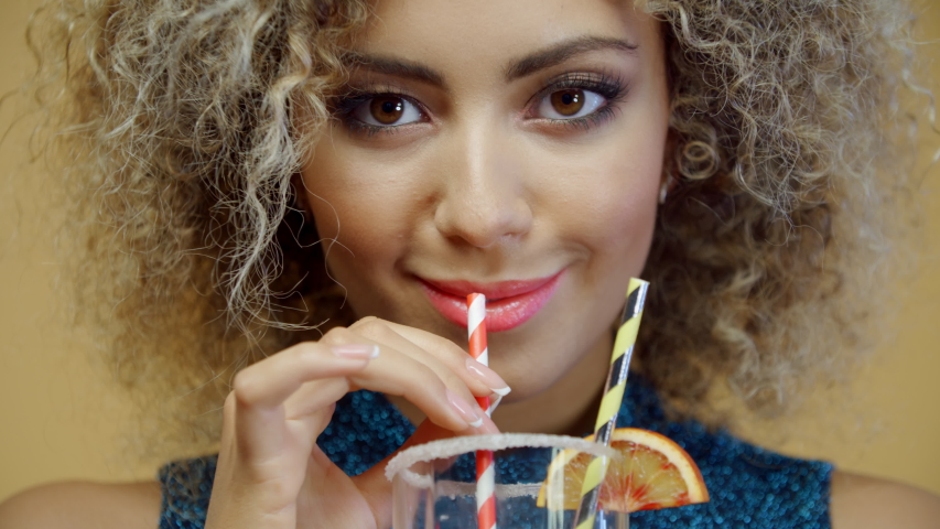 Close-up portrait beautiful mixed race woman drinks orange cocktail through paper straw on yellow background. Joyful hispanic female enjoys holding glass of refreshing drink and smiling