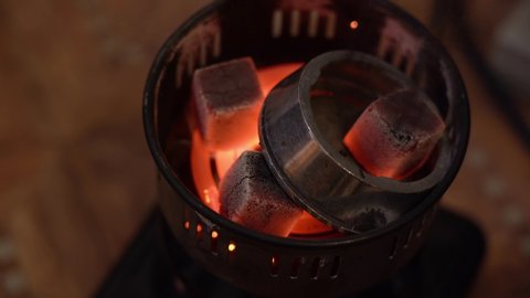 A man hookahman with metal tongs puts hot smouldering coals in kaloud on a special portable electric spiral stove for ignition close up. Top View. The concept of a hookah for hiking.