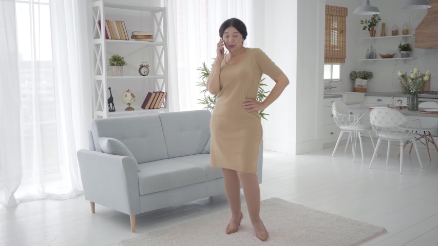 Cute senior Caucasian woman talking on the phone, putting device on couch and dancing indoors. Portrait of cheerful middle-aged lady in elegant beige dress enjoying retirement at home.