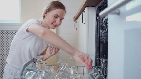 A woman opens the dishwasher in her kitchen and pulls out glasses