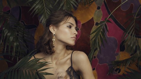 Tanned beautiful girl model with large gold earrings on a motley background among tropical leaves.