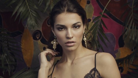 Tanned beautiful girl model with large gold earrings on a motley background among tropical leaves.