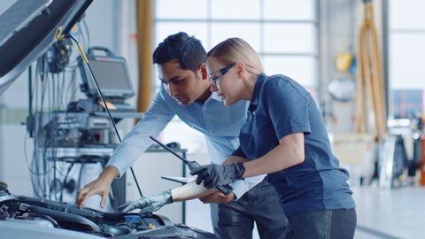 Manager Checks Tasts on a Tablet Computer and Explains an Engine Breakdown to an Female Mechanic. Car Service Employees Inspect Car's Engine Bay with a LED Lamp. Modern Clean Workshop.