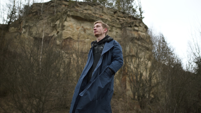 Handsome Young Guy Standing Near Rock. Stylish Redheaded Boy With Beard And Ear Piercing. Guy In Coat. Wind Around Him.