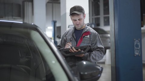 Portrait of serious Caucasian maintenance engineer checking automobile with digital equipment. Adult man in workwear using computer to verify car damages. Repair shop, service station.