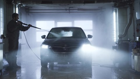 Adult confident Caucasian man washing vehicle in auto care shop. Professional washer cleaning car with manual high-pressure water sprayer. Service, maintenance, automotive industry.