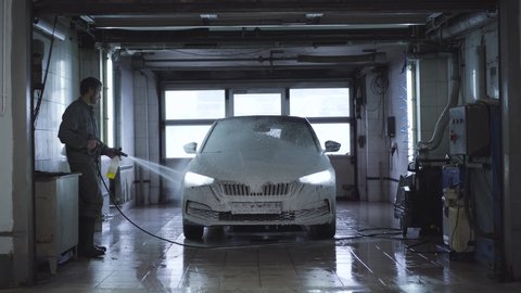 Wide shot of worker washing vehicle in car wash. Caucasian employee using high-pressure washer at service station. Maintenance of automobile, automotive industry.
