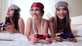 Three young girls playing competitive video game with joysticks, enjoying online game. Female friends in sleeping masks lying in bed in morning, sunshine. Electronic sports, entertainment concept.