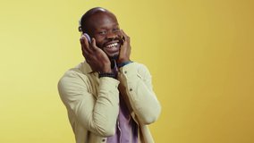 Happy emotional african man listening to favorite music via headphones smile cheerful enjoy sounfs rhythm posing at ywlloe wall background.