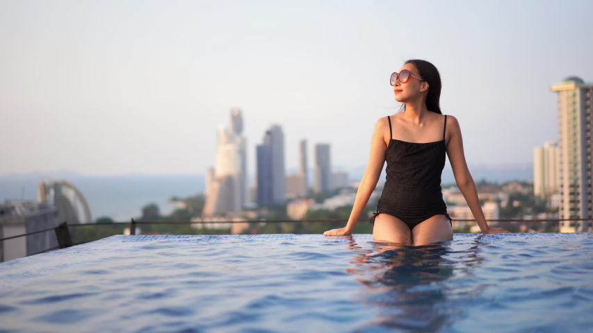 Pretty woman wearing a black bathing suit and sunglasses sitting on the edge of a rippling infinity pool with a breeze blowing through her long brunette hair and a cityscape in the background