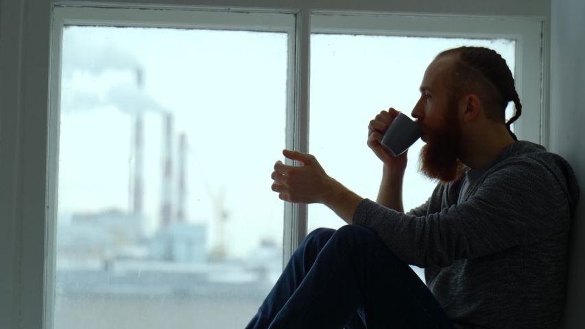 Portrait of a red-haired man with a beard sitting by the window against the background of pipes emitting smoke. | Shutterstock HD Video #1047673957
