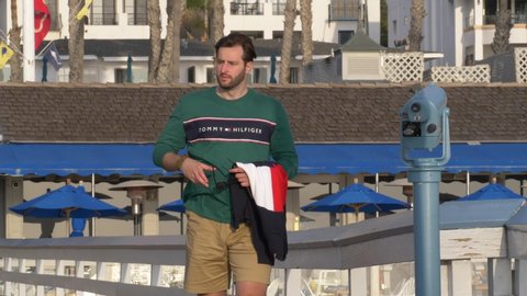A handsome, young guy, a man in tommy shirt , shorts and glasses is walking along in walking at hte pier. San Clemente USA January 16th 2020