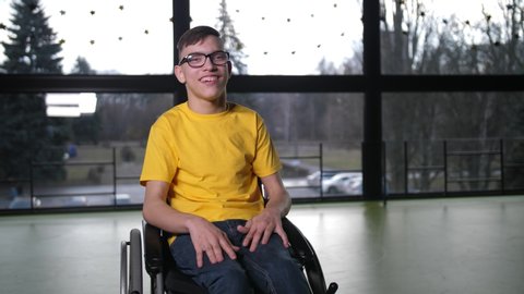 Close-up portrait of happy boy with cerebral palsy sitting in wheelchair in the middle of sport hall. Positive disabled teenager in eyeglasses and bright yellow T-shirt smiling at camera