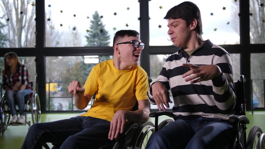 Close-up of positive teen boys with cerebral palsy having fun talking while sitting in wheelchairs next to each other, two disabled women communicating on background by window
