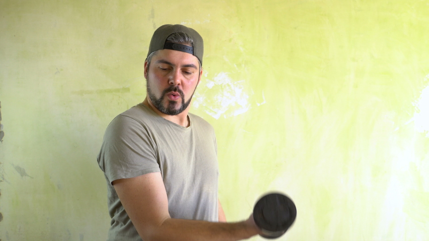 A man in a cap with a beard is standing against the wall. Training with dumbbells. Fitness exercise. A positive look. Joyful face. Military T-shirt.