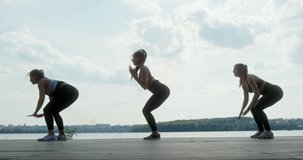 Three athletic young women working out together on a wooden deck overlooking water leaping, stretching and squatting in a side view against a high key sky in a health and fitness concept