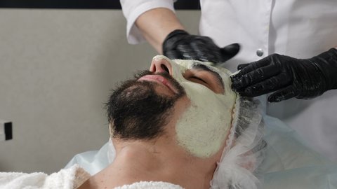 Concept of beauty salon and spa for men. Beautician applying facial cosmetic mask massaging with gloved hands. Spa therapy for handsome man getting rejuvenating facial treatment. 4 k footage