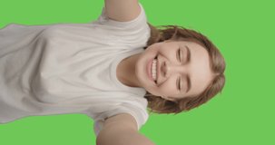 Young smiling hipster blond woman in white t-shirt posing over green screen background. Girl taking selfie self portrait photos on smartphone. Vertical 4k raw video footage slow motion 60 fps