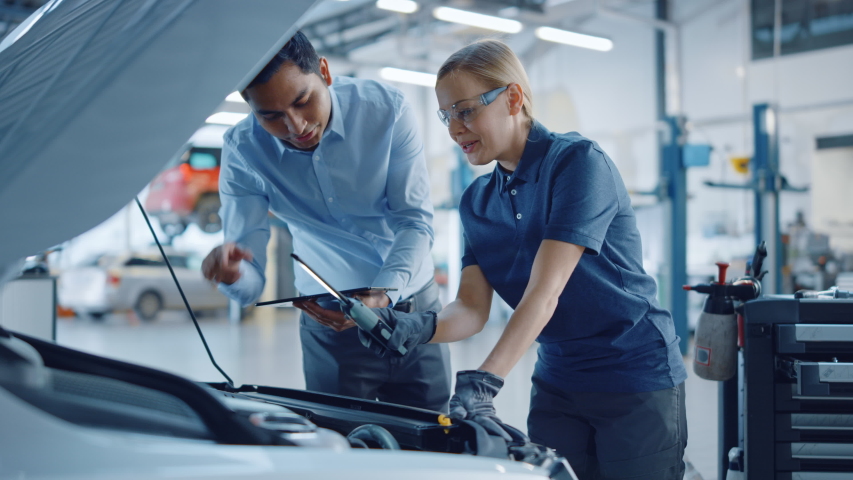 Instructor with a Tablet Computer is Giving a Task for a Future Mechanic. Female Student Inspects the Car Engine. Assistant is Checking the Cause of a Breakdown in the Vehicle in a Car Service. Royalty-Free Stock Footage #1047697183