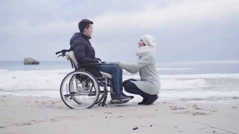 Young woman sitting on hunkers in front of boy in wheelchair and talking. Mother or invalid tender supporting handicapped young man on sea shore or river bank outdoors. Joy, lifestyle, help.