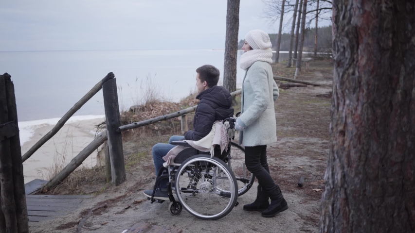 Side view of cheerful Caucasian boy with a disability talking withtender and laughing. Portrait of positive young men with disabilities man using a wheelchair enjoying leisure outdoors. Lifestyle- care- help.