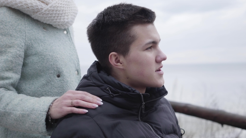 Close-up portrait of young brunette Caucasian boy in wheelchair talking with unrecognizable adult woman on river bank. Smiling disabled man looking back and away outdoors. Disability, paralysis.
