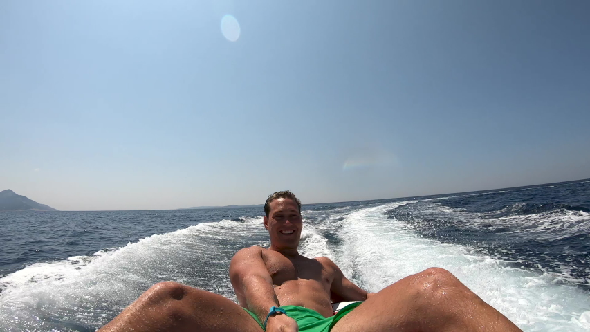 Man smiling and sitting on an inflatable ring towed by the boat, filming himself with go-pro, making happy faces into the camera.  Royalty-Free Stock Footage #1047703657