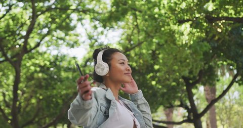 Low angle side view of a happy mixed race woman with long dark hair out and about in the city streets during the day, walking with headphones on, listening to music, holding a smartphone, smiling with Video Stok