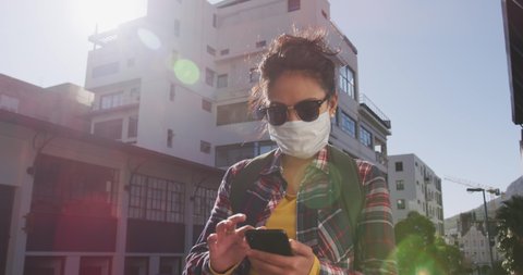Low angle front view of a mixed race woman with long dark hair out and about in the city streets during the day, wearing sunglasses and a face mask against air pollution and Coronavirus Covid19