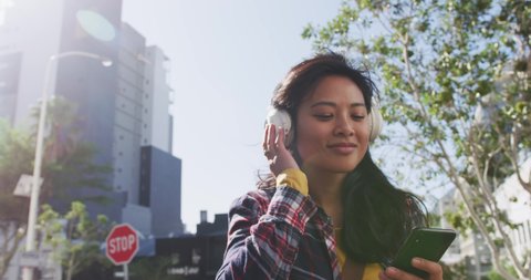 Low angle front view of a happy mixed race woman with long dark hair out and about in the city streets during the day, walking with headphones on, listening to music, holding a smartphone, smiling