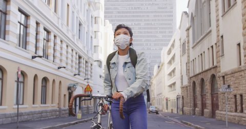 Front view of a mixed race woman with long dark hair out and about in the city streets during the day, wearing a face mask against air pollution and coronavirus Covid19, walking with her bicycle