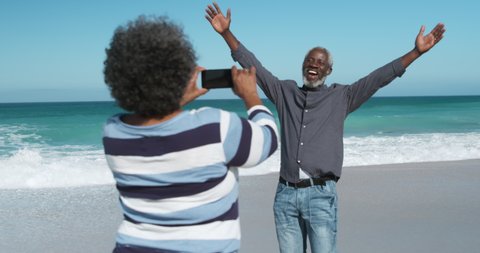 Front view of a senior African American couple standing on the beach with blue sky and sea in the background, the woman taking photos with smartphone, the man dancing and smiling, in slow motion