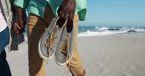 Side view of mid section of a senior African American couple enjoying time on the beach with blue sky and sea in the background, holding hands and shoes in slow motion 库存视频