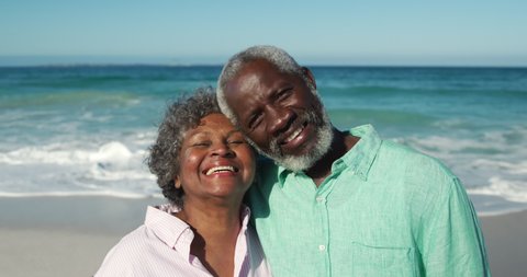 Portrait of a senior African American couple standing on the beach with blue sky and sea in the background, embracing each other, looking at camera and smiling in slow motion