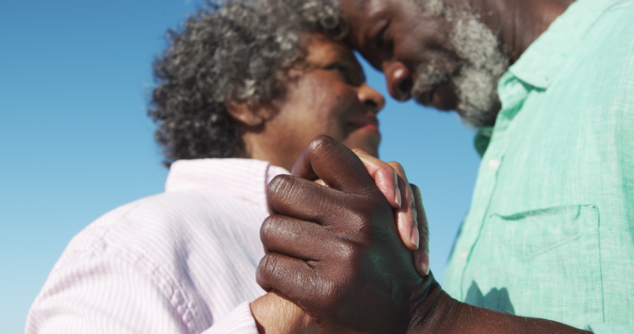 Low angle side view close up of a senior African American couple standing on the beach with blue sky in the background, holding hands, dancing and having fun in slow motion Royalty-Free Stock Footage #1047710398