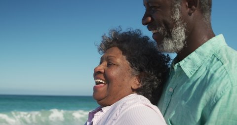 Low angle side view close up of a happy senior African American couple in love standing on the beach with blue sky and sea in the background, embracing each other and smiling in slow motion