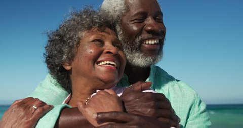 Front view close up of a happy senior African American couple standing on the beach with blue sky and sea in the background, embracing each other and smiling in slow motion