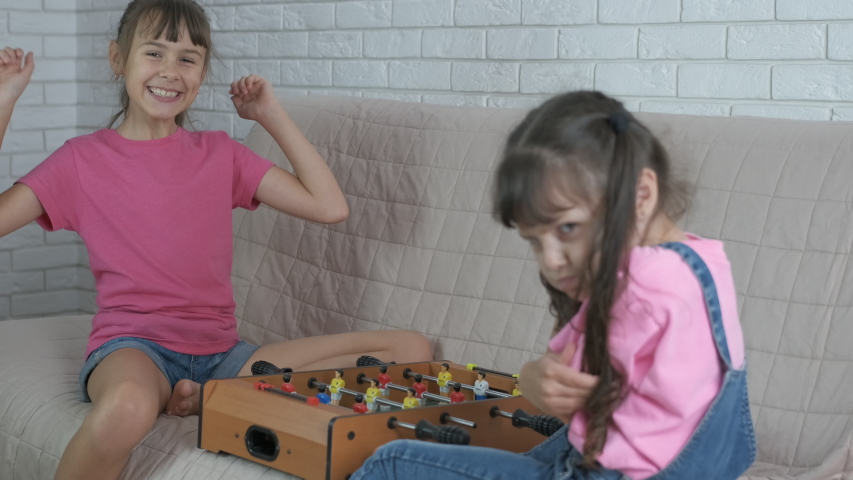 Happy sisters play table football in the room. One child win the game.