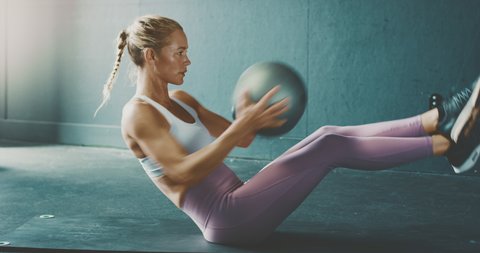 Fit attractive blonde woman doing core abdominal workout exercises with a medicine ball, determined woman training in the gym to achieve her fitness goals